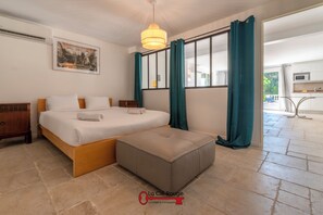 Ein modernes Schlafzimmer mit einem Bett, einem hölzernen Nachttisch, einem grauen Pouf, einer Pendelleuchte und einem großen Fenster mit türkisfarbenen Vorhängen.