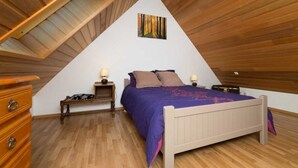 Ein Schlafzimmer mit schrägen Wänden, einem Bett mit violetter Bettdecke, Holznachttischen und einer Kommode.