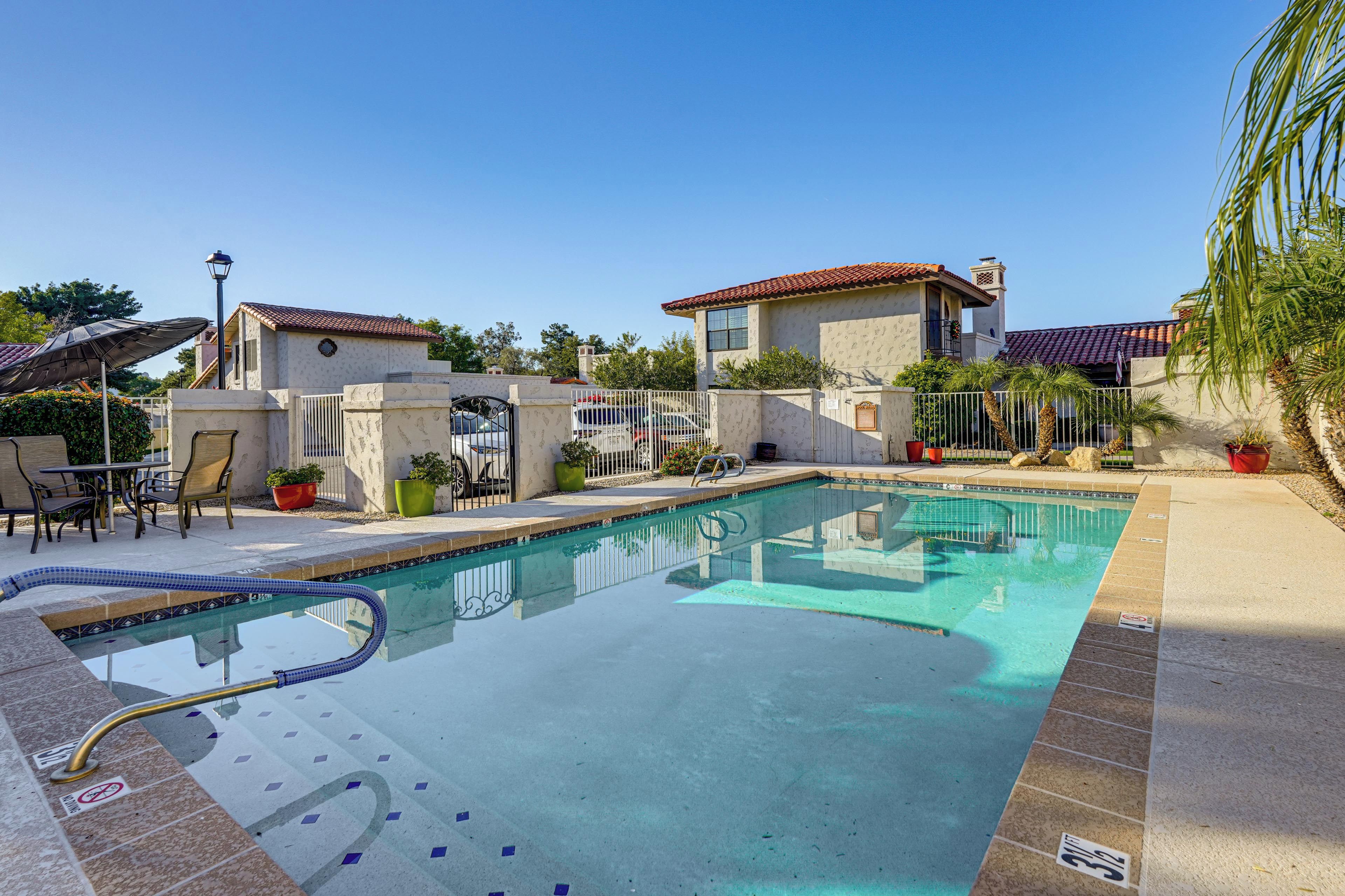 A swimming pool with a tiled deck, a covered seating area, and a building with a red-tiled roof in the background.