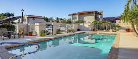 Una piscina con solárium empedrado, un área de estar techada y un edificio con techo de tejas rojas al fondo.
