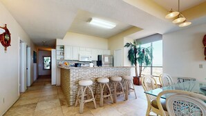 A modern kitchen with white cabinets, a central island with stools, and a dining area with a glass table and wicker chairs. - Shorefront Corner PH, 3br: Suntide III - Unit 1201 (South Padre Island)