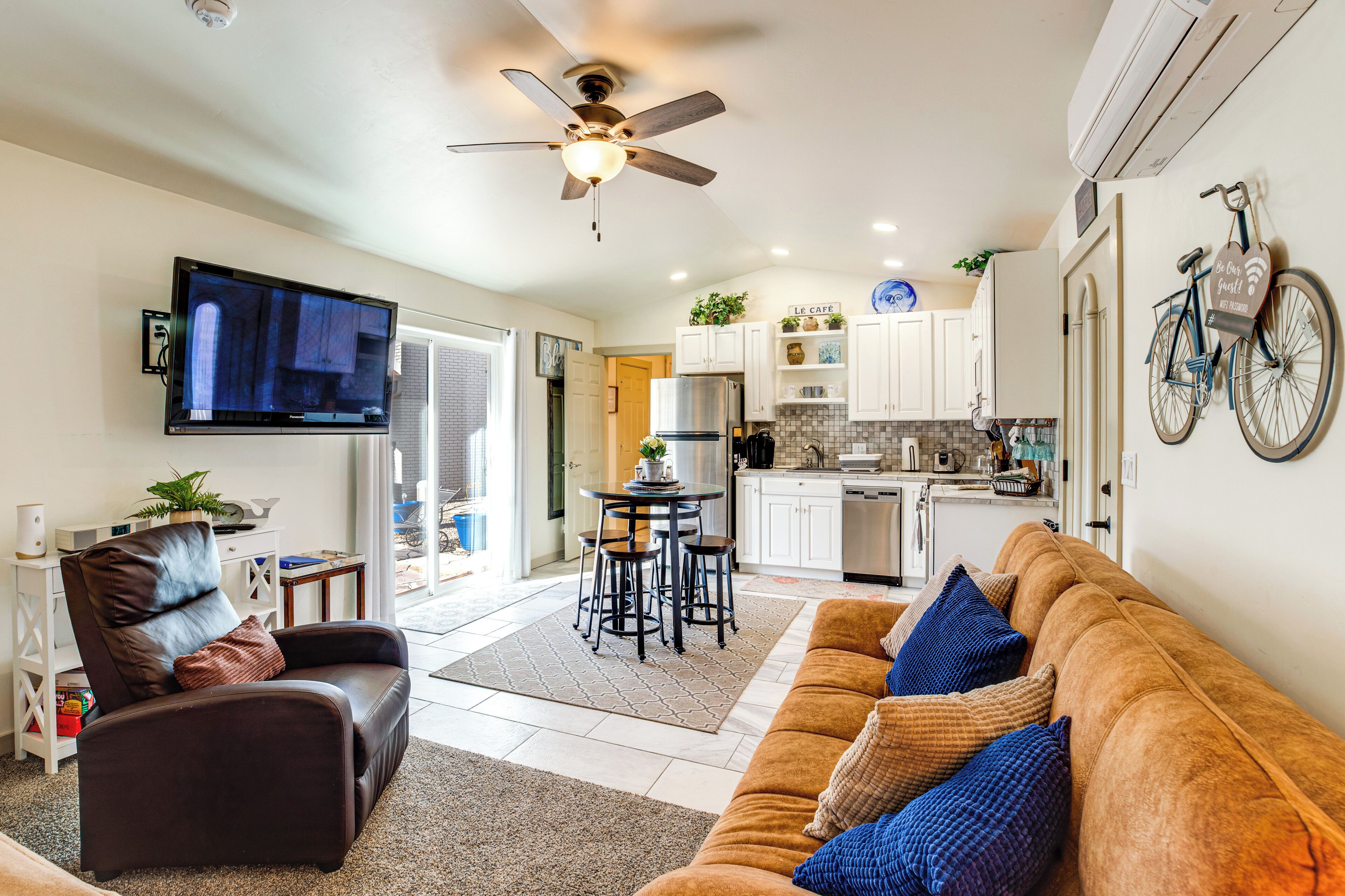 A modern living room with a sofa, a television, a ceiling fan, and a kitchen area with white cabinets and a bicycle mounted on the wall.