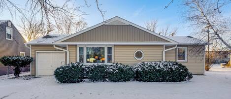 A residential house with a garage, a snow-covered lawn, and leafless trees.