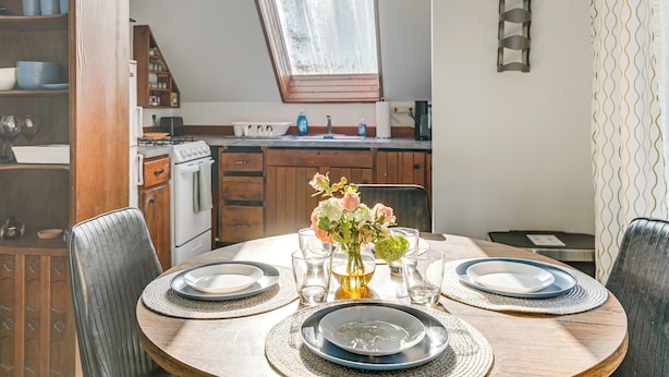 Un coin repas avec une table ronde dressée pour quatre personnes, un vase rempli de fleurs et, au fond, une cuisine.