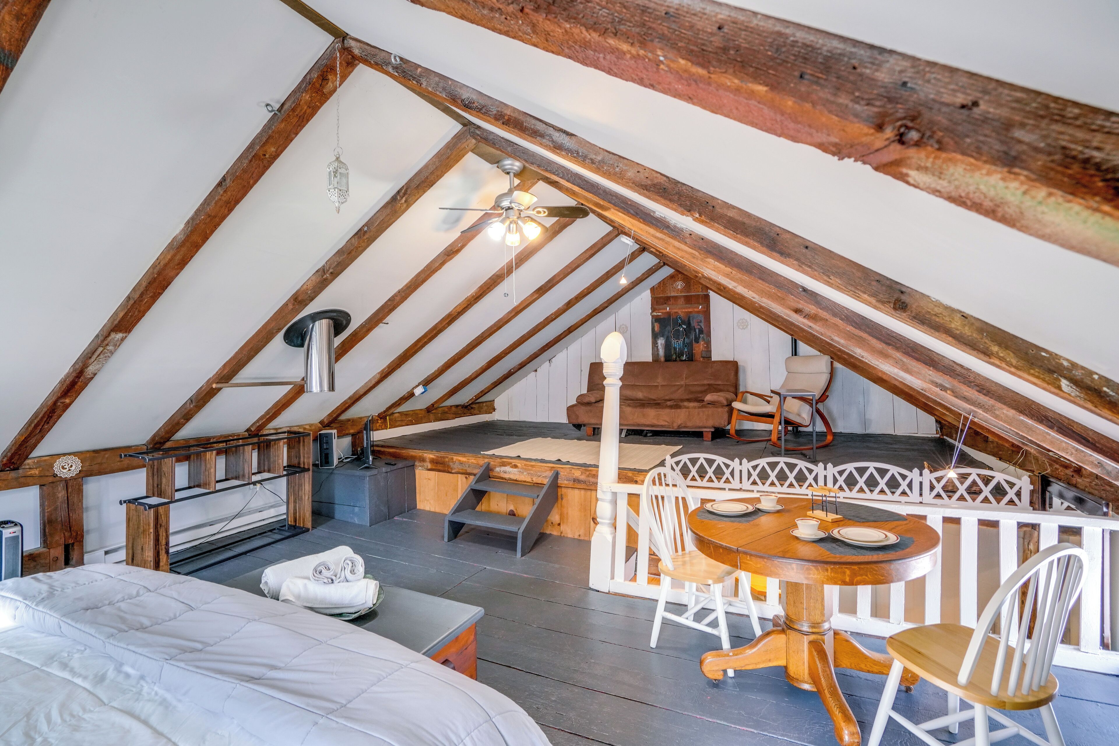 A cozy attic bedroom with a wooden ceiling, a bed, a small table with chairs, and a white fence.