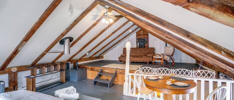 A cozy attic bedroom with a wooden ceiling, a bed, a small table with chairs, and a white fence.