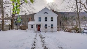 A white two-story house with a red door, surrounded by snow-covered ground and trees. - Hike, Fish & Ski: Riverside Barn Loft in Lee (South Lee)