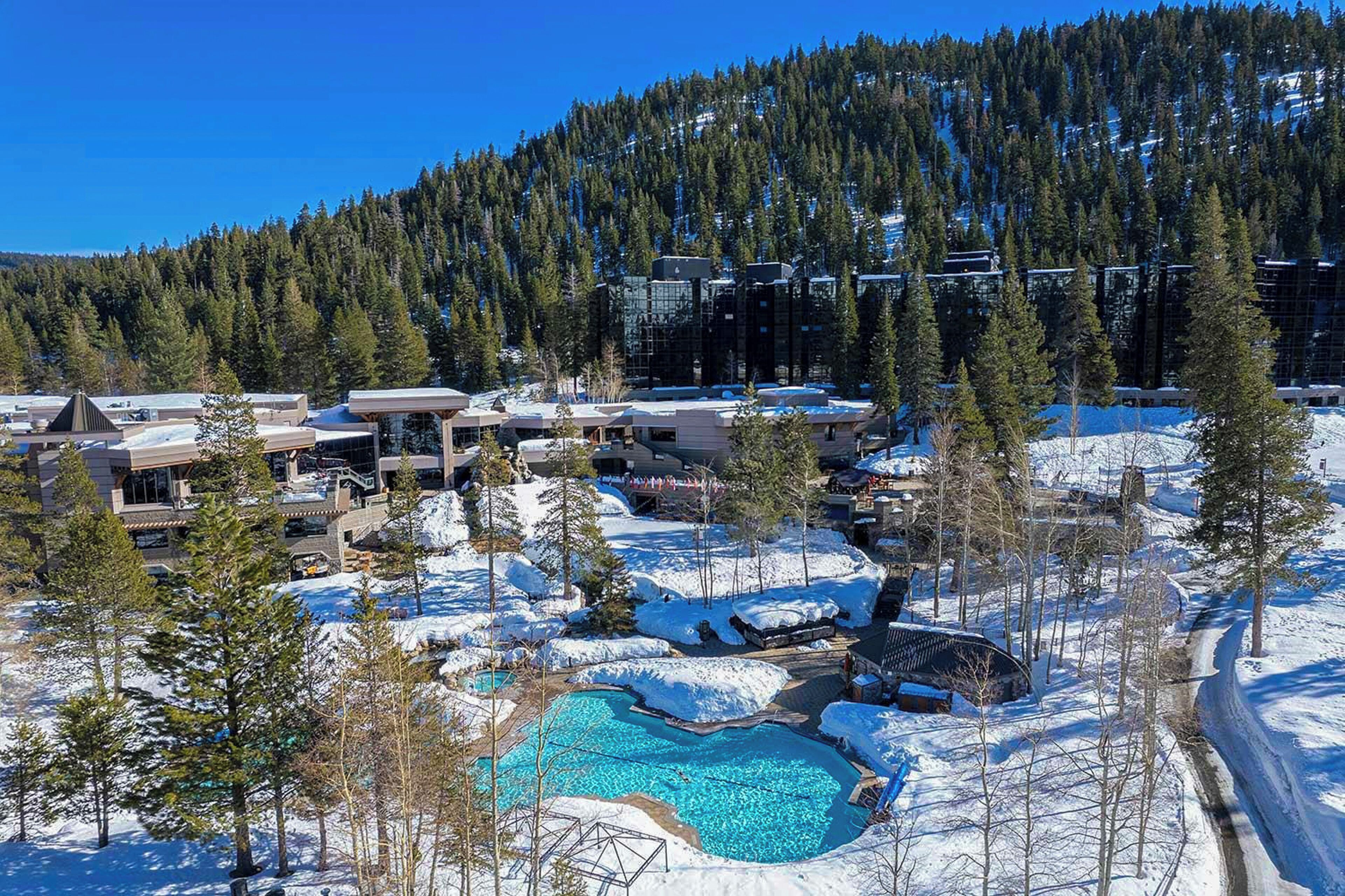 Un resort con piscina rodeado de nieve, edificios y una montaña boscosa al fondo.
