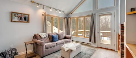 A spacious living room with a high ceiling, large windows, a ceiling fan, and a cozy seating area with a sofa and ottoman.