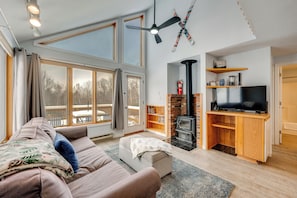 A living room with a sofa, ottoman, and a TV on a wooden stand. There is a fireplace, bookshelves, and a large window with curtains. - Sled, Ski & Hike! Mtn-view Condo in Thornton (Thornton)