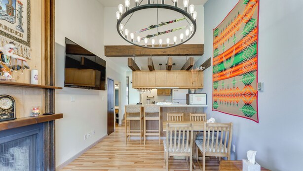 A modern living room with a fireplace, wooden flooring, a dining area with chairs, a kitchen with wooden cabinets, and a large decorative wall hanging. - Walk to Angel Fire Resort: Pet-friendly Getaway (Angel Fire)
