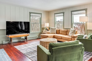 A living room with a sectional sofa, a television, a lamp, and a large window with blinds. - Dine & Unwind! Overlook Home at Rabbit Hill Inn (St. Johnsbury)