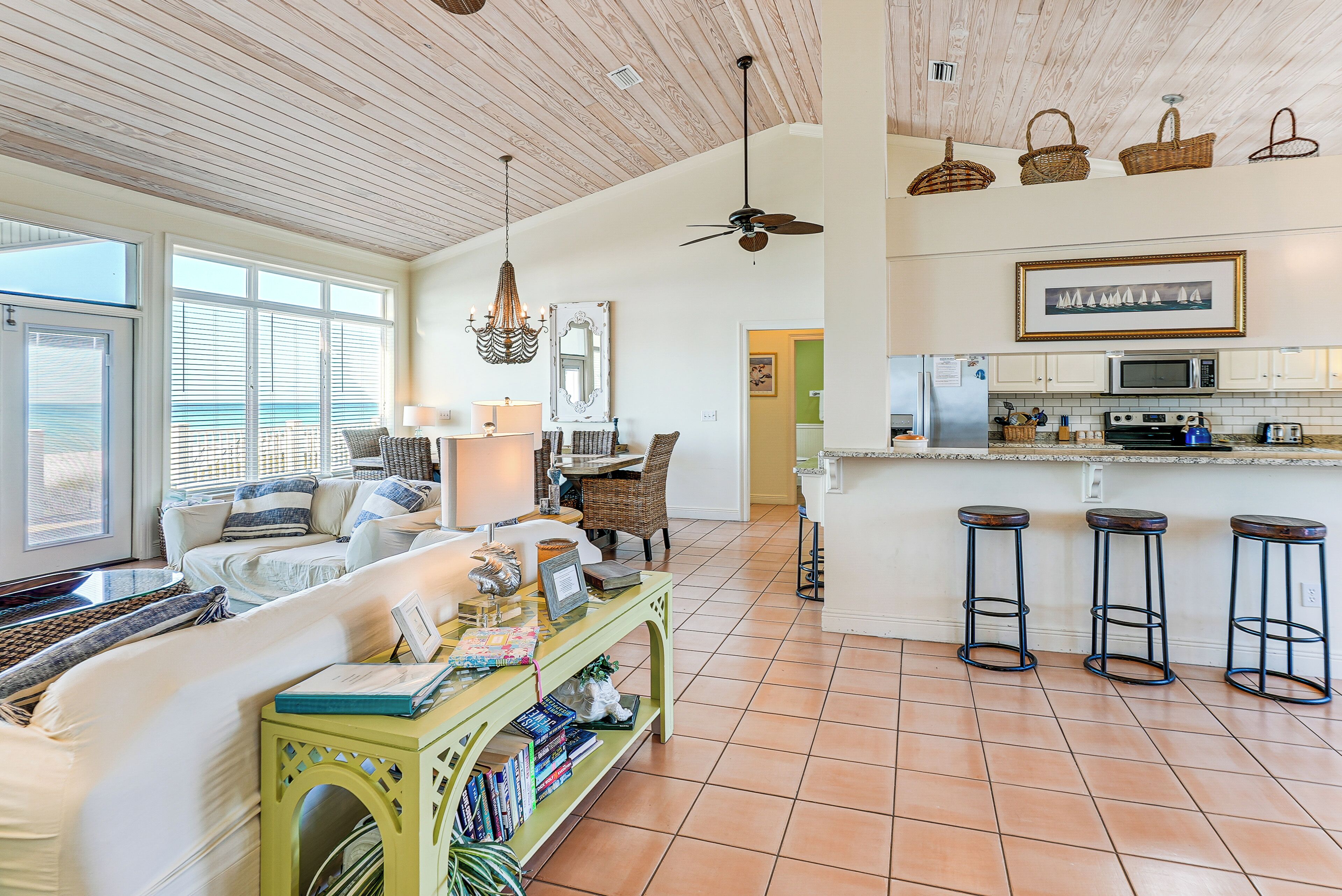 Un salón amplio con un ventanal grande, una consola verde y una zona de cocina con microondas y taburetes.