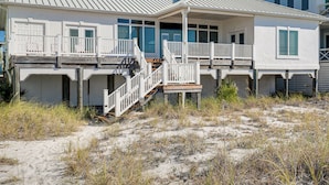 Een huis aan het strand met een overdekte veranda, een trap naar de hoofdingang en een zandstrand op de voorgrond.
