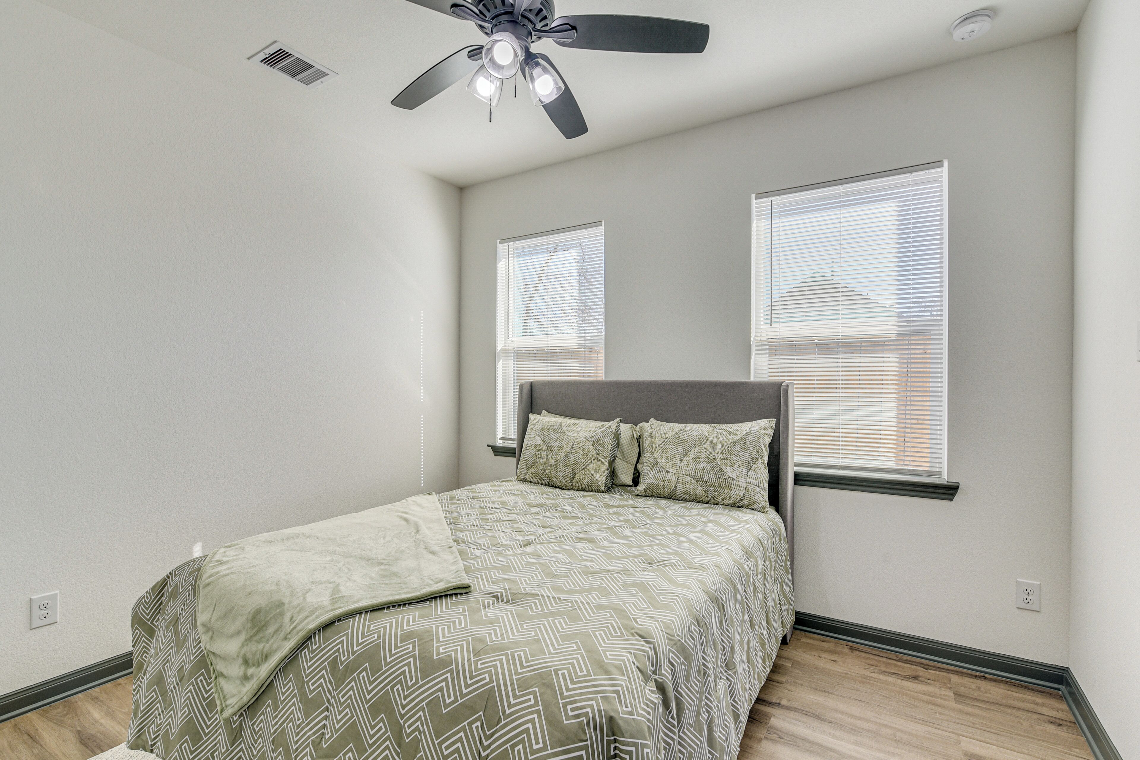 A bedroom with a bed, ceiling fan, two windows with blinds, and a wooden floor.