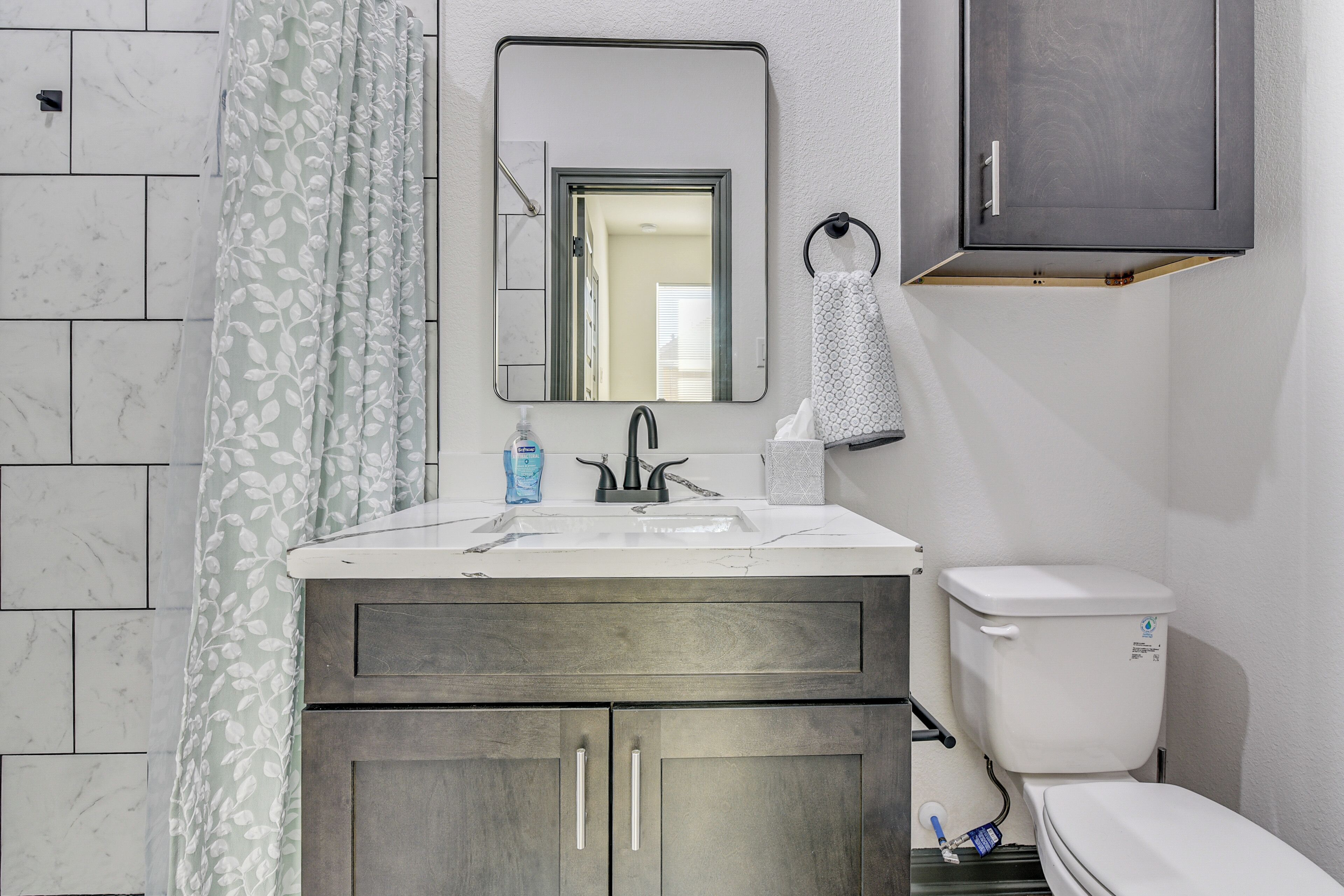 Une salle de bain avec un lavabo blanc, des meubles en bois foncé, des toilettes et un rideau de douche orné d’un motif de feuilles.