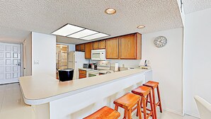 A kitchen with wooden cabinets, a white countertop, and a built-in microwave. There are four wooden stools and a clock on the wall. - Shorefront 2br, Luxe Perks: Suntide III - Unit 704 (South Padre Island)