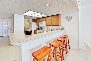 Une cuisine avec des armoires en bois, un plan de travail blanc et un four à micro-ondes encastré. On y trouve quatre tabourets en bois et une horloge au mur.