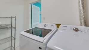 Een wasserette met twee witte Samsung wasmachines, een metalen droogrek en een deur naar een kamer met een blauwgroene wand.