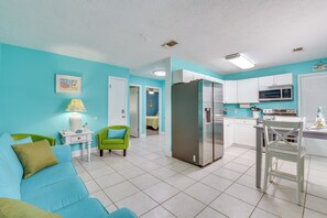 A modern kitchen with white cabinets, a stainless steel refrigerator, and a microwave. There is a dining table with chairs and a green armchair. The walls are painted teal, and there is a framed picture on the wall. - Walk to Beach & Rick Seltzer Park! PCB Getaway (Panama City Beach)