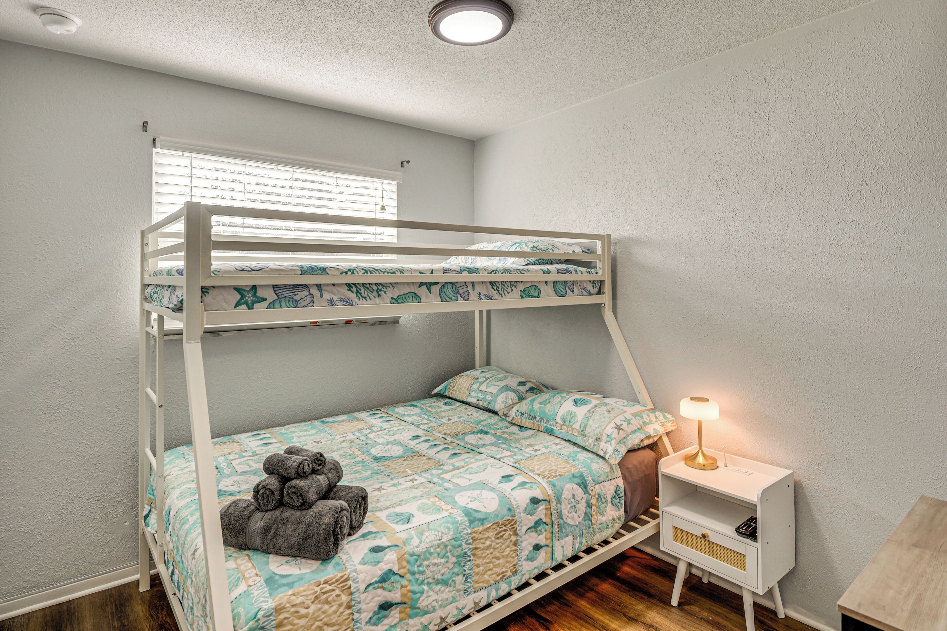 Un lit superposé avec une courtepointe, une table de chevet avec une lampe et une fenêtre avec des stores.