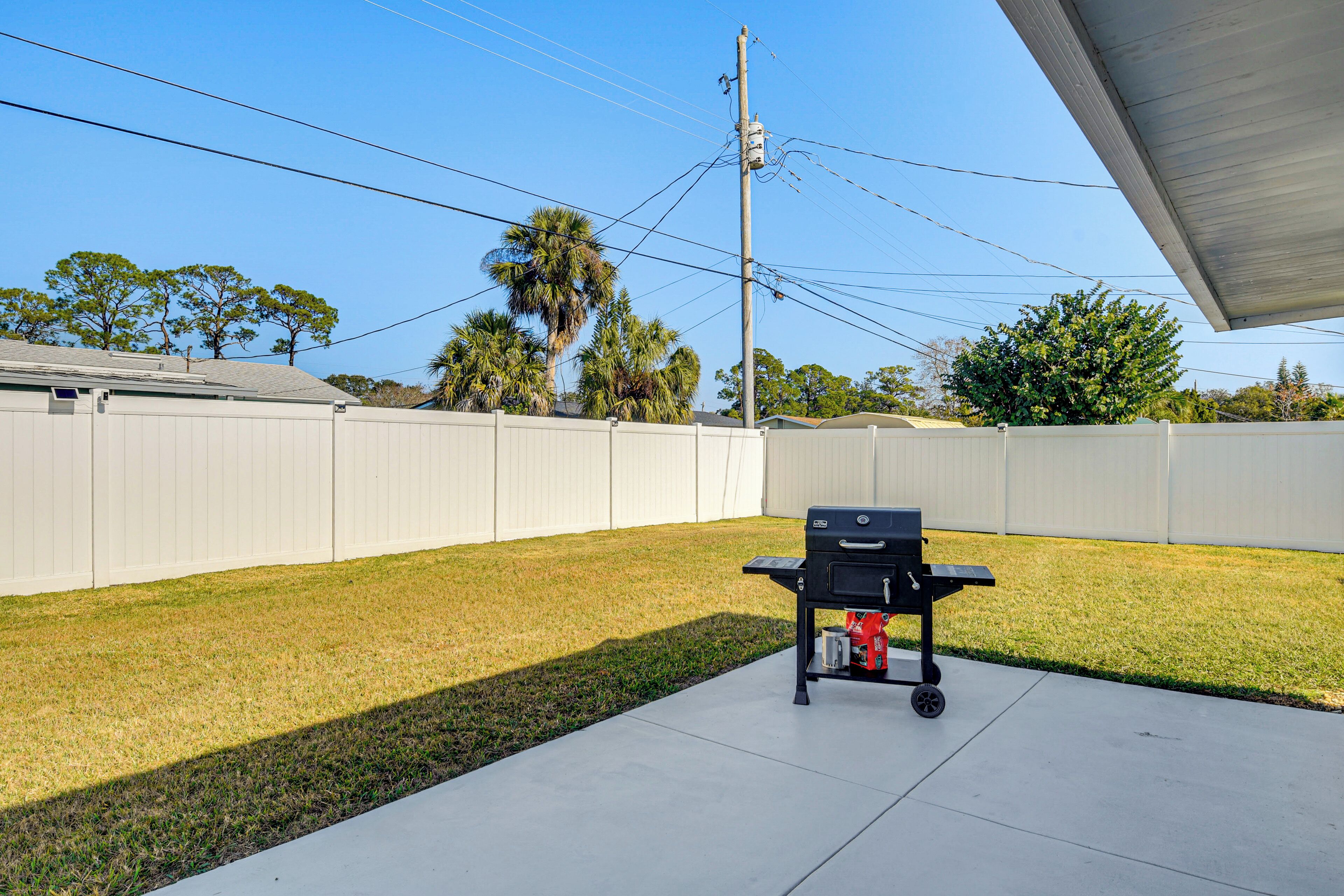 A backyard with a barbecue grill, a concrete patio, a white fence, and trees in the background.