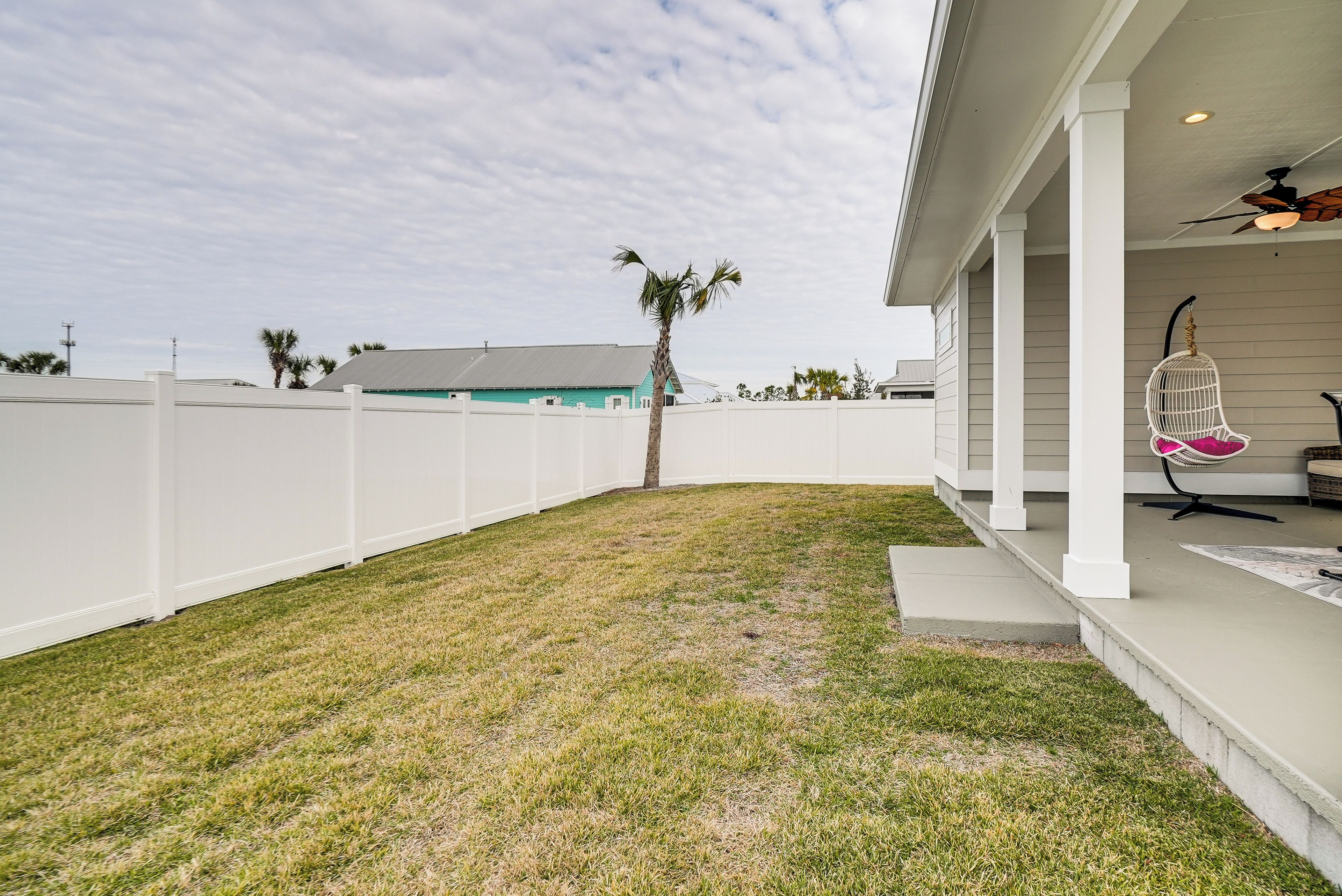 A backyard with a white fence, a palm tree, and a porch with a hanging chair.