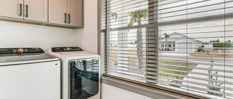 A laundry room with a washer and dryer, wooden flooring, a window with blinds, and a view of a residential area.