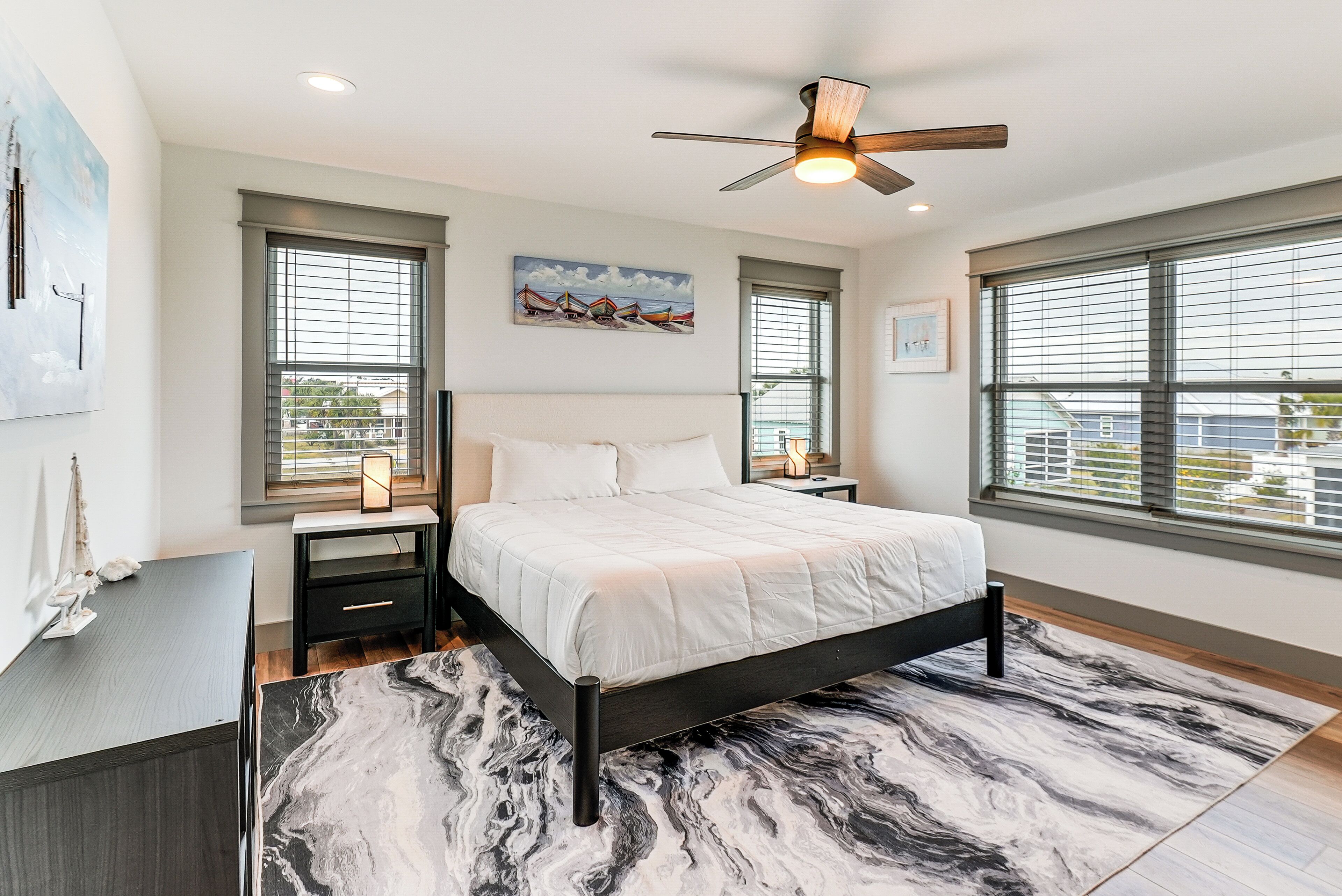 A modern bedroom with a large bed, a ceiling fan, and large windows with blinds.