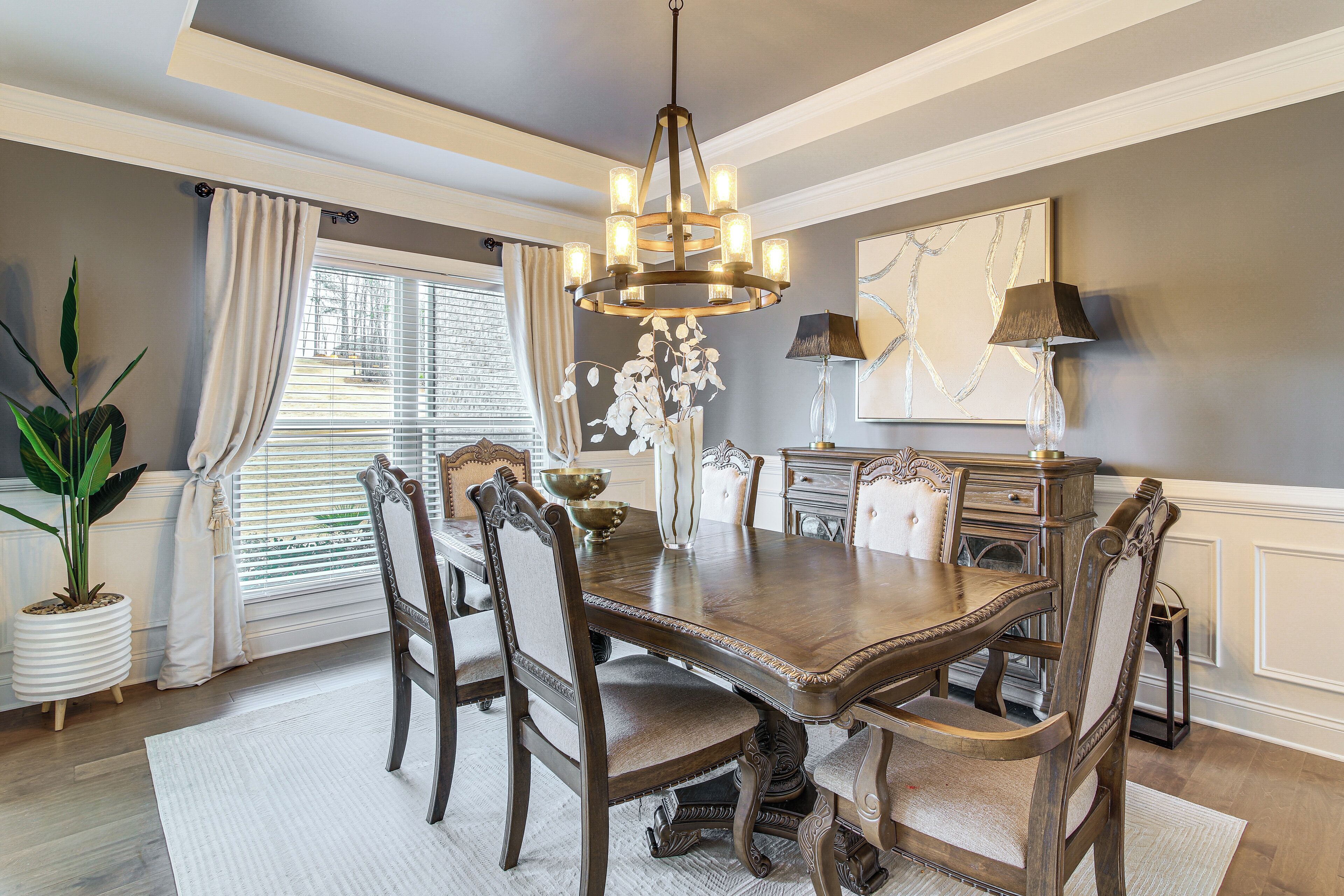 A dining room with a wooden table, six chairs, a chandelier, and a large window with curtains.