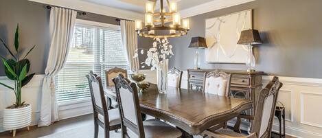 A dining room with a wooden table, six chairs, a chandelier, and a large window with curtains.