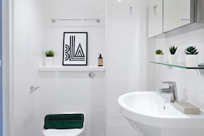 Une salle de bain moderne avec un lavabo blanc, une œuvre d’art encadrée au-dessus du miroir et de petites plantes en pot sur l’étagère.