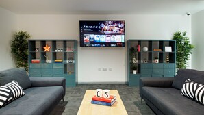 A modern living room with two grey sofas, a coffee table with books, a TV displaying 'Friends', and green shelving units with decorative items. - For Students Only Ensuites in Southampton (Southampton)