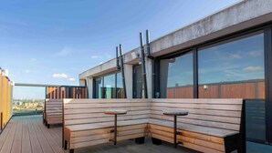 En tagterrasse med træbænke og borde, store vinduer og udsigt over byen.