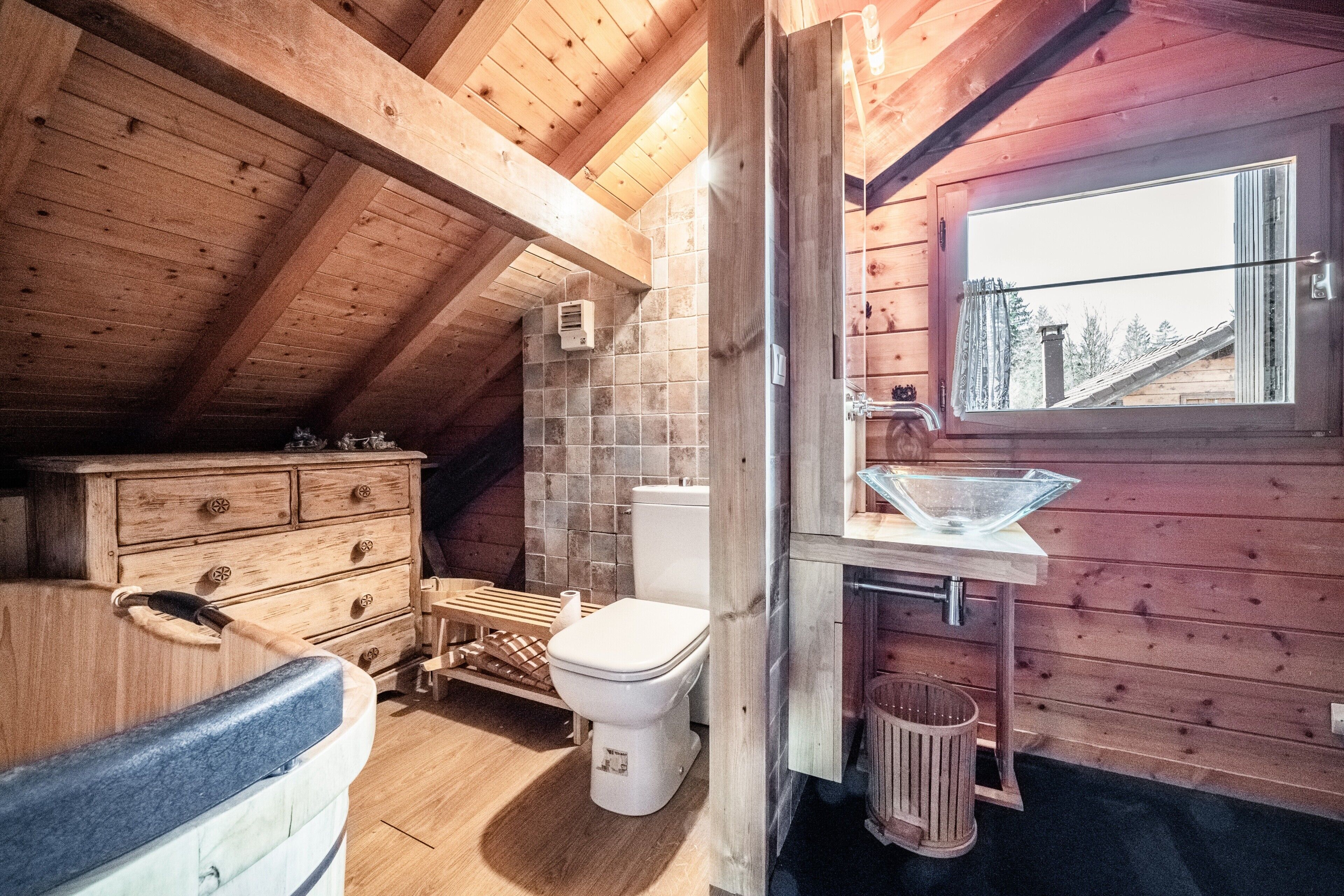 A wooden cabin interior with a wooden ceiling, a window with a view, a wooden dresser, a toilet, and a sink with a glass basin.