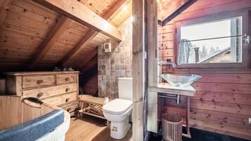 A wooden cabin interior with a wooden ceiling, a window with a view, a wooden dresser, a toilet, and a sink with a glass basin.