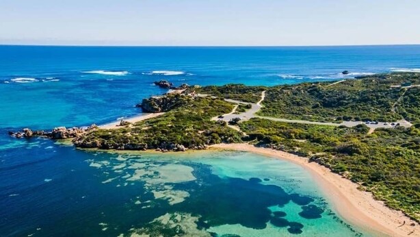 Vista aerea di una zona costiera con acque blu cristalline, una spiaggia sabbiosa e un paesaggio collinare dalla vegetazione rada.