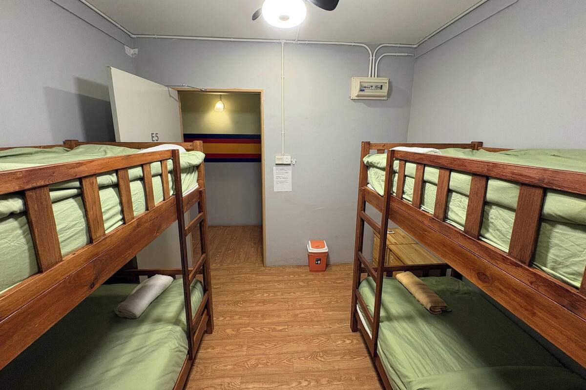 A dormitory room with bunk beds, a ceiling fan, a wall-mounted electrical box, and a small trash bin.
