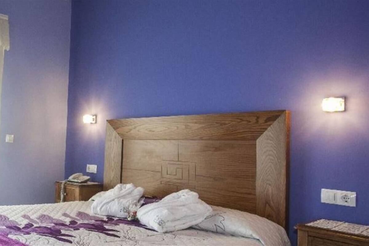 A bedroom with a wooden headboard, a bed with white linens and a purple throw, a nightstand with a phone, and a window with white curtains.