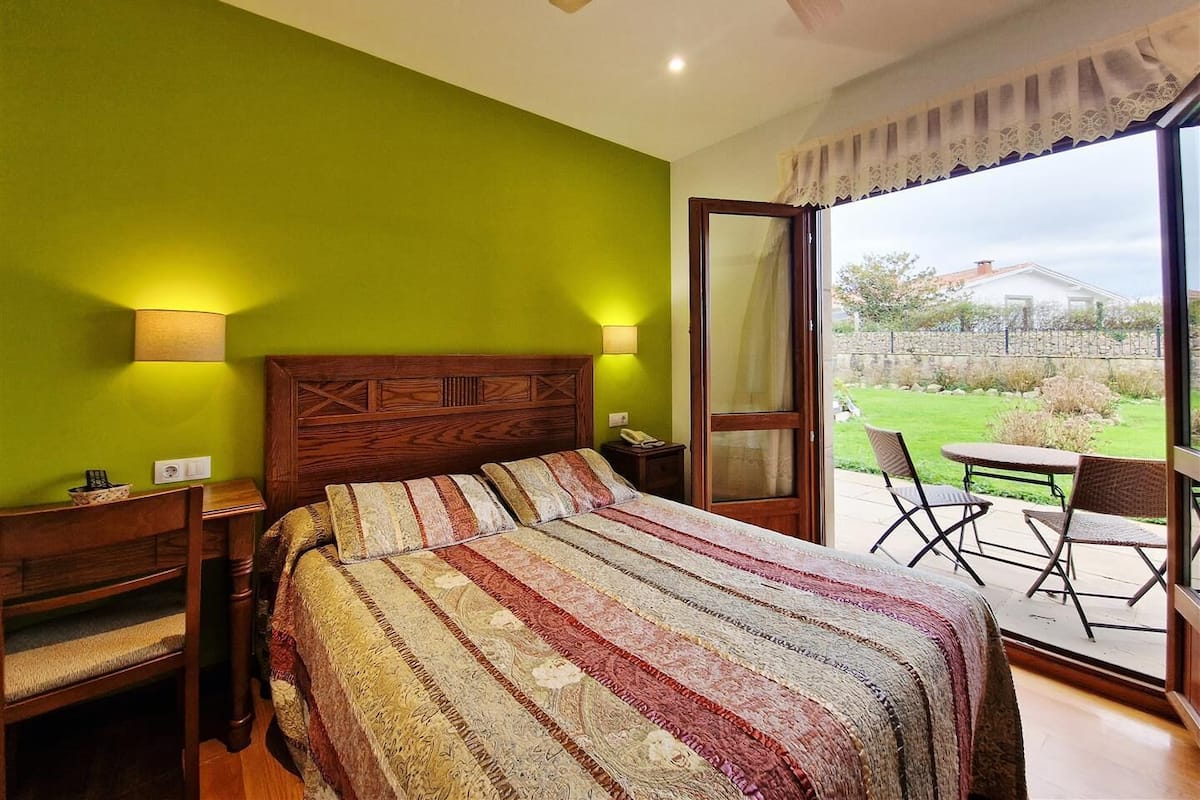 A bedroom with a large bed, a wooden headboard, two bedside lamps, a nightstand, and a view of a garden through a sliding glass door.