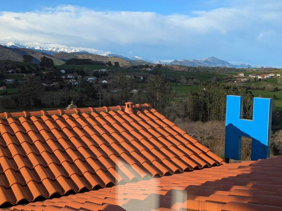 Ein mit Terrakotta-Ziegeln gedecktes Dach mit einem auffälligen, blauen 'H'-Schild, das auf eine gebirgige Landschaft mit verstreuten Häusern blickt.