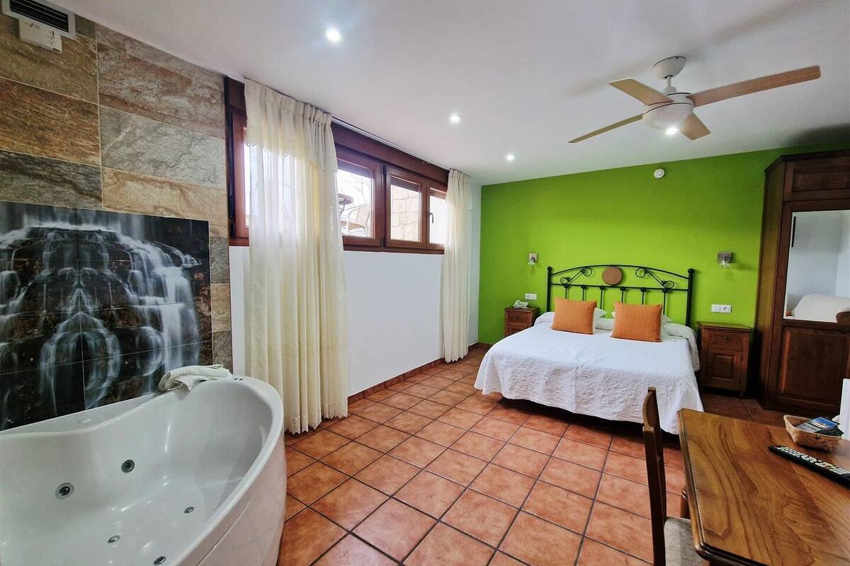 A room with a large bathtub, a bed with two pillows, a ceiling fan, and a green wall.