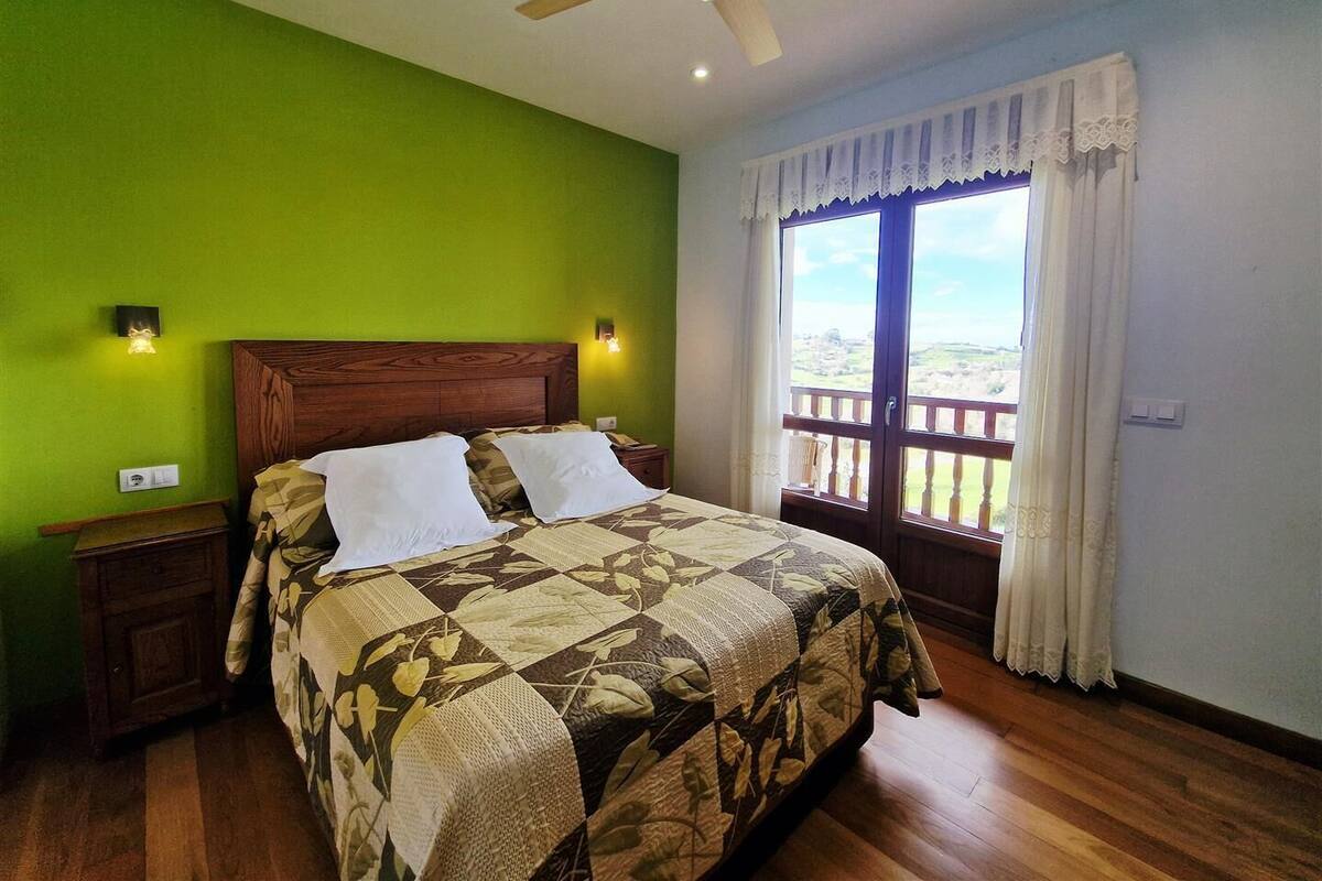 A bedroom with a wooden bed, green walls, a balcony with a view, and a ceiling fan.