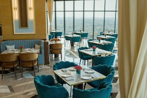 A modern restaurant with teal chairs and tables, a city view through large windows, and a mix of seating arrangements.