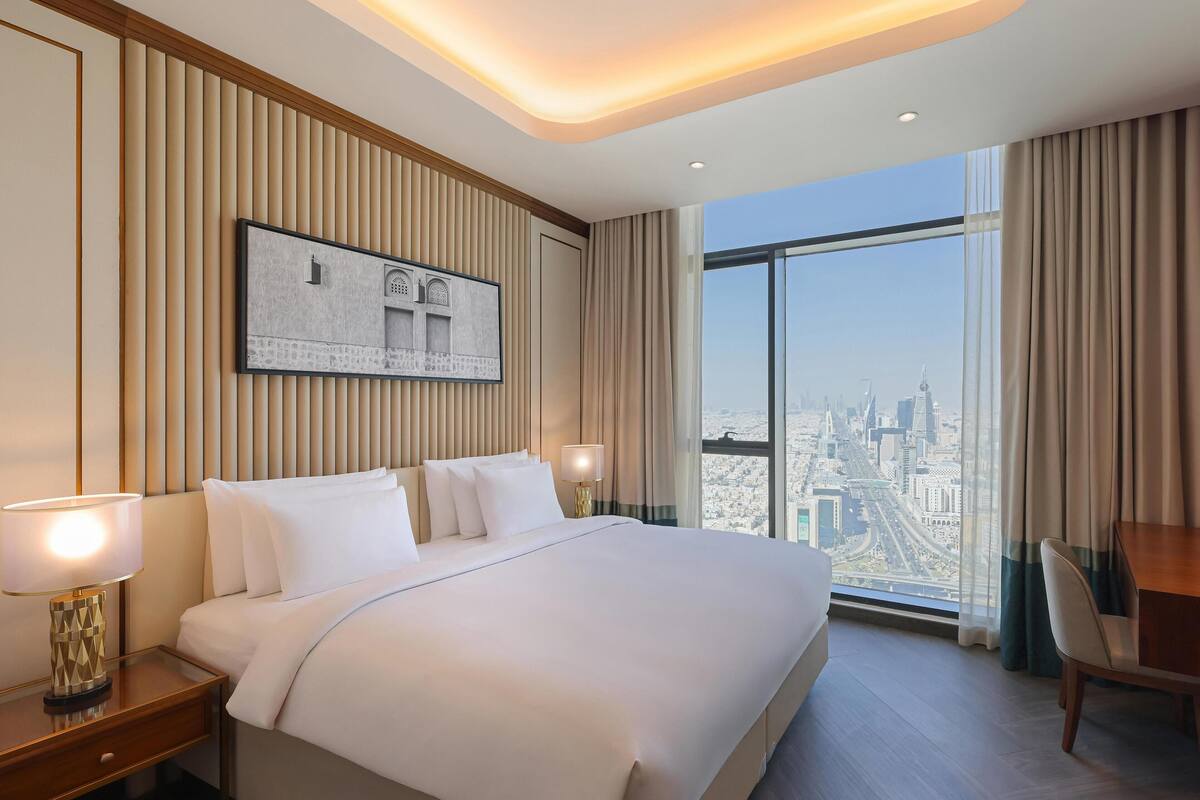 A hotel room with a large bed, a nightstand with a lamp, a framed picture on the wall, and a city view through the window.