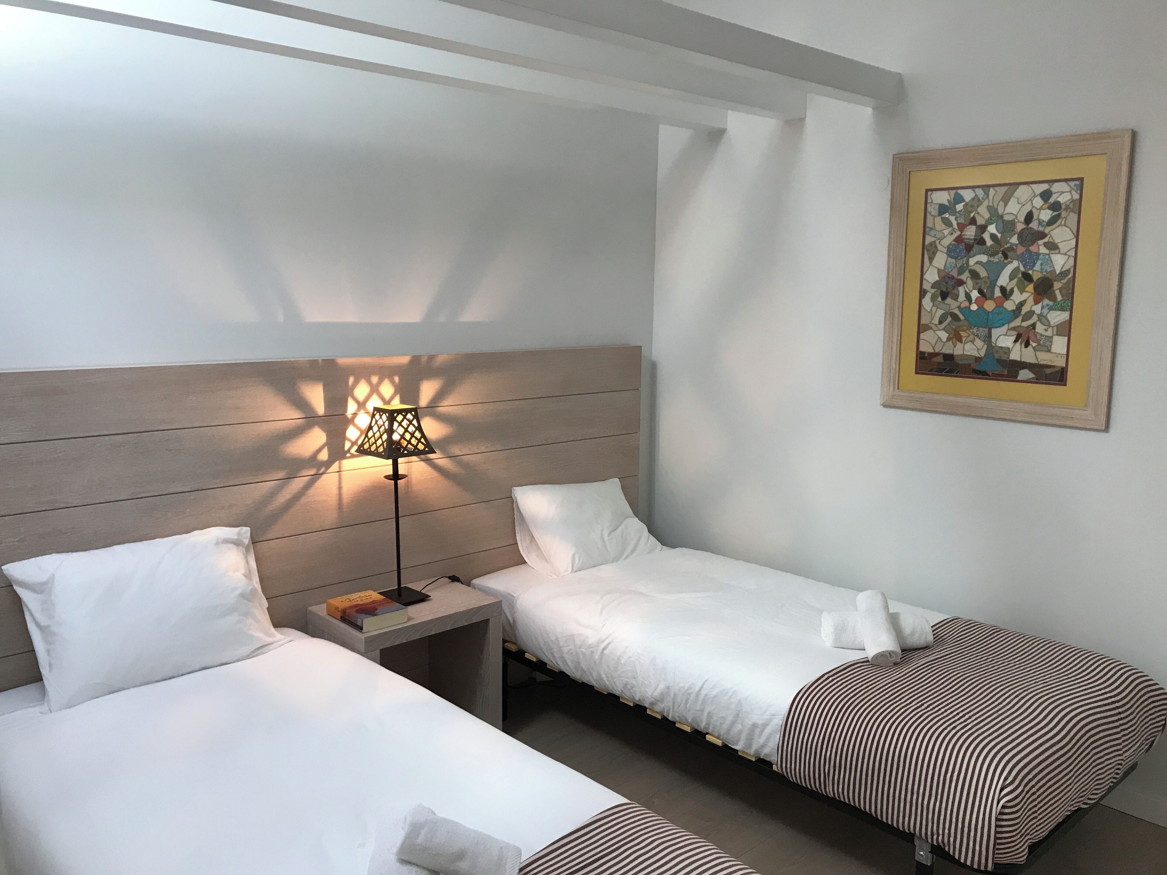 A hotel room with two beds, a nightstand with a lamp, a painting on the wall, and a wooden headboard.