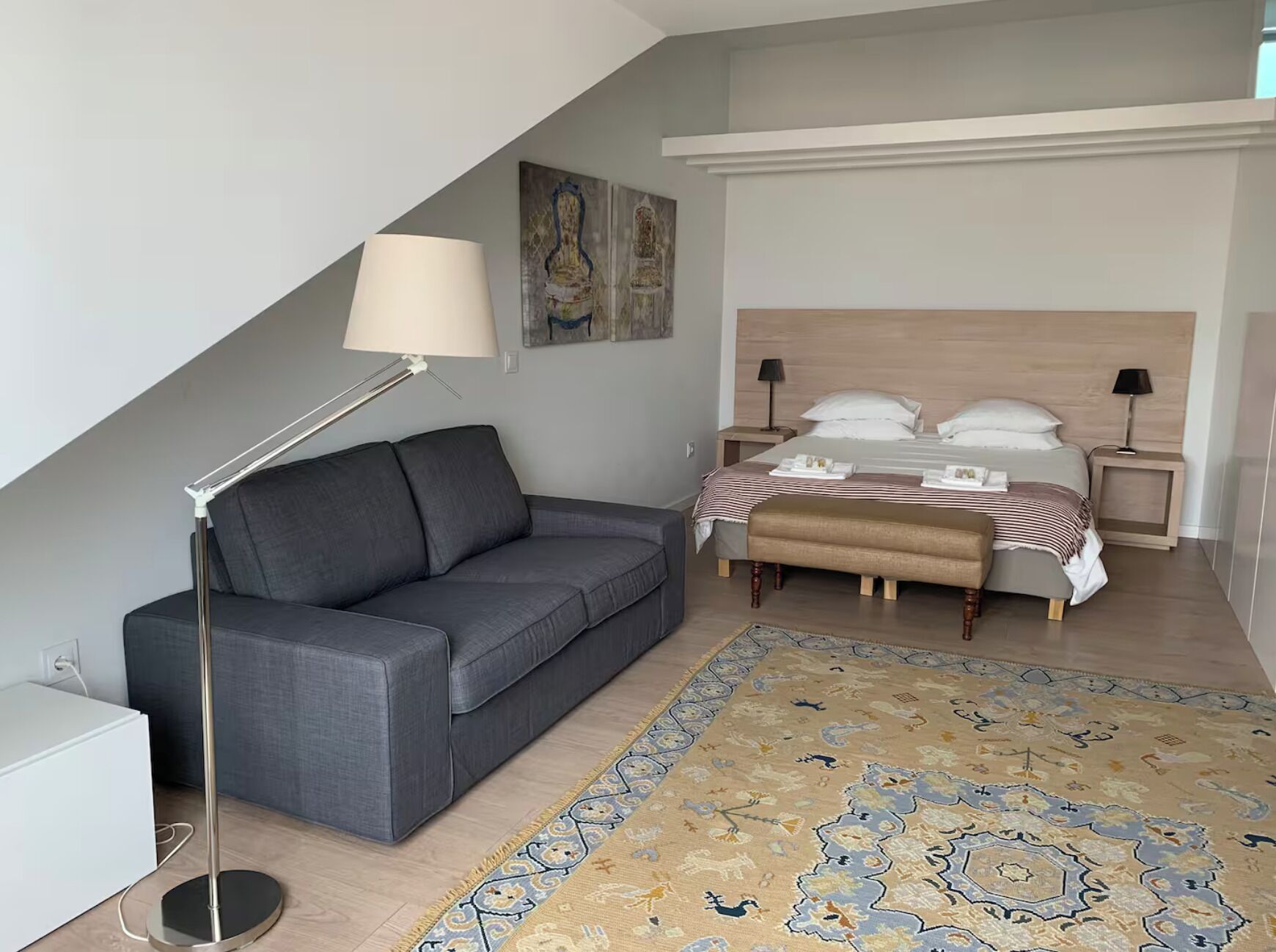 Un dormitorio moderno con un sofá gris, una cama de madera con colcha a rayas, una alfombra estampada y un cuadro en la pared.