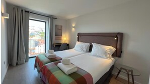 Habitació d'hotel amb dos llits, un escriptori i vista a edificis a través d'una finestra gran.