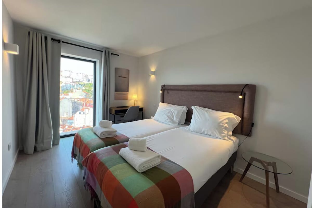 Habitació d'hotel amb dos llits, un escriptori i vista a edificis a través d'una finestra gran.
