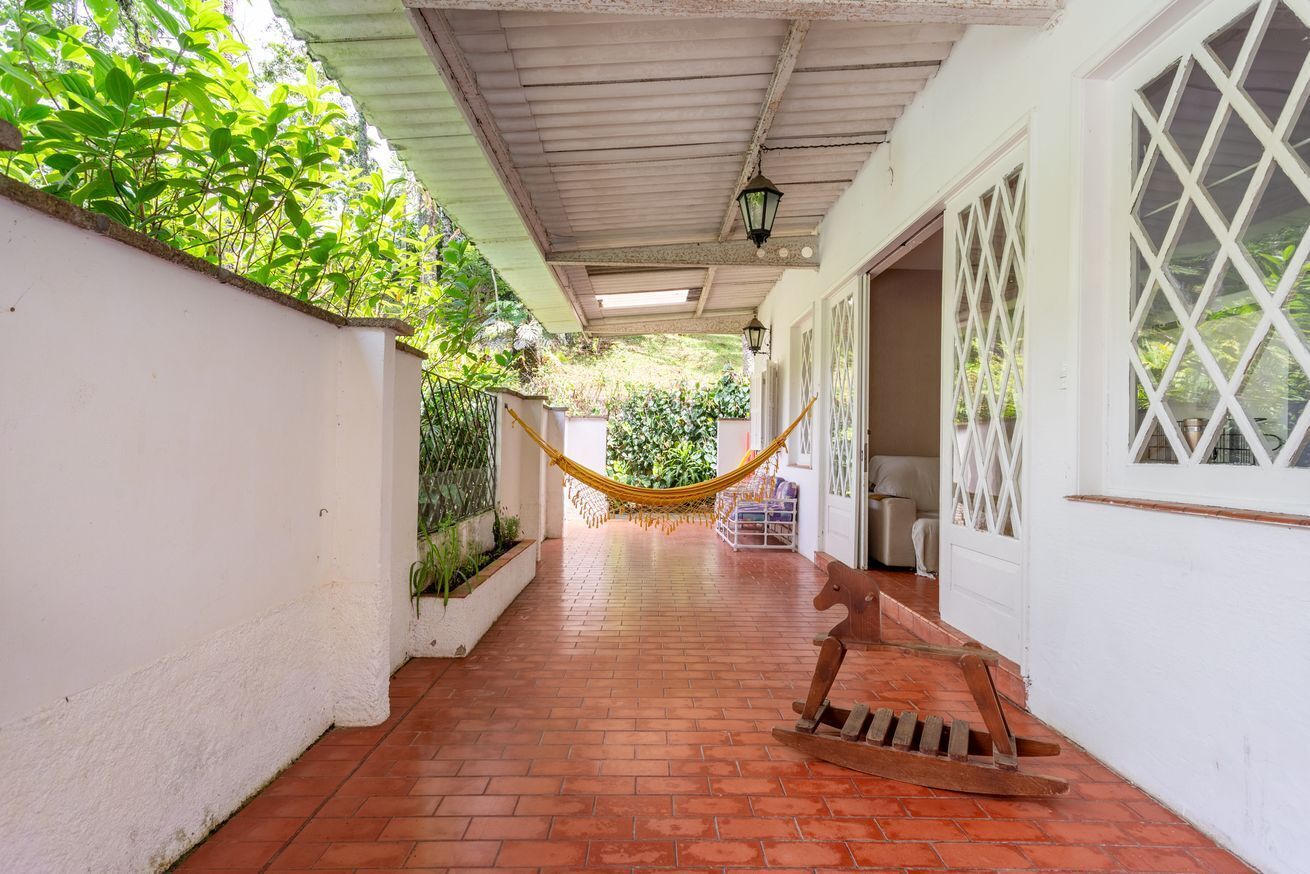 A covered outdoor corridor with a hammock, a wooden rocking chair, and a white wall with lattice windows.